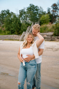 A couple stands on a sandy beach; the man embraces the woman from behind while she rests her arm on her head. Both are casually dressed in white tops and jeans, with greenery in the background.