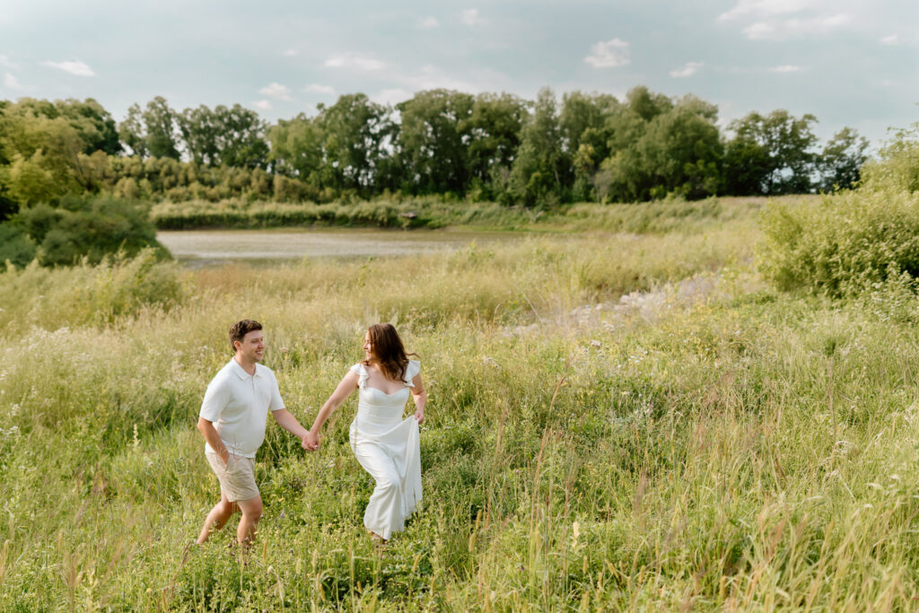 Couple walking through grassy field near pond during Miss Millie’s Flower Farm engagement session in Manitoba.