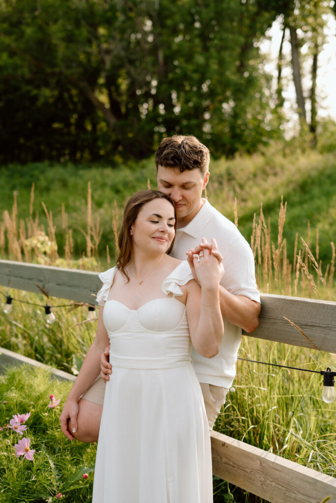 Couple embracing beside a wooden fence during a summer engagement session at Miss Millie’s Flower Farm in Manitoba with warm afternoon light and greenery.